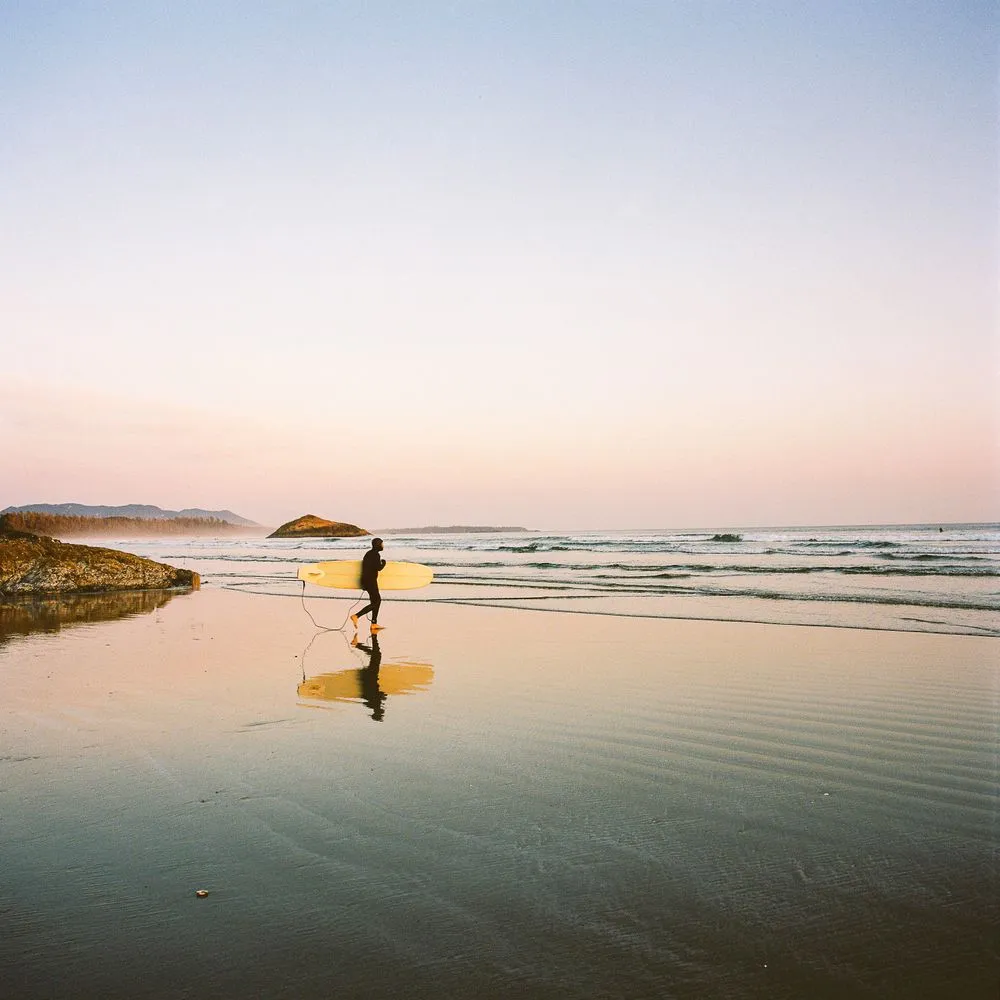 Surfer walking up a reflective beach carrying a yellow board