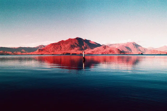 Red-tinted mountain reflected in calm water, with a small figure out in the shallows