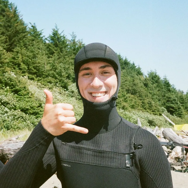 Andrew on the beach in a hooded wetsuit, grinning and throwing a shaka